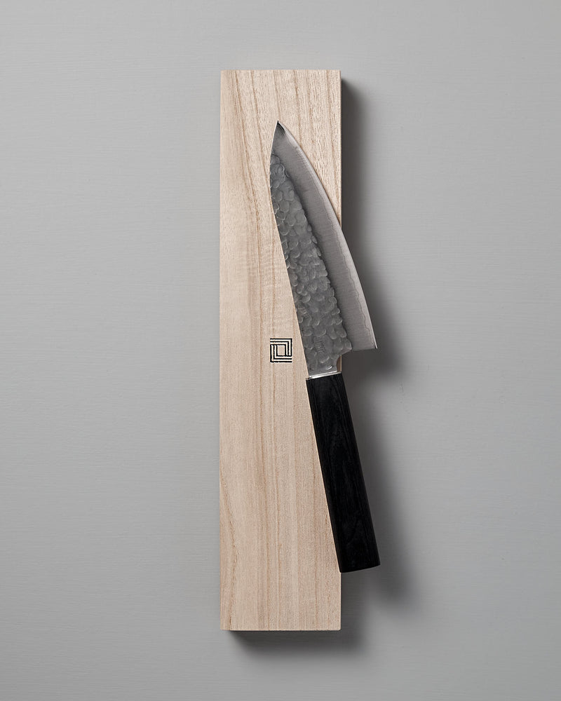 The Yamato Deba Knife by Shizu Hamono Co., featuring a Sanmai steel blade with a hammered finish, rests on a wooden block against a gray background.