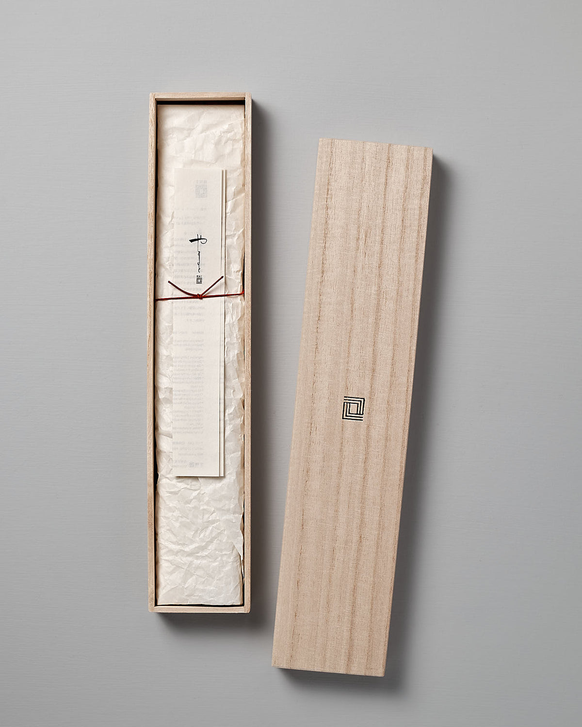 A rectangular wooden box with its lid removed, revealing a document and a piece of textured white paper inside. The items are sealed with a red string and feature minimalistic text and designs, reminiscent of the elegant craftsmanship found in Shizu Hamono Co.'s Yamato Deba Knife.