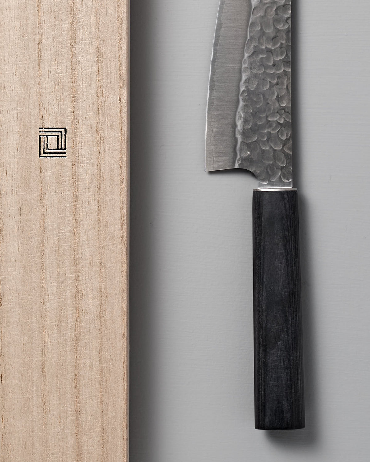 Close-up of a Shizu Hamono Co. Yamato Deba Knife featuring a textured Sanmai steel blade and dark handle, placed beside a wooden cutting board adorned with a minimalist square logo.