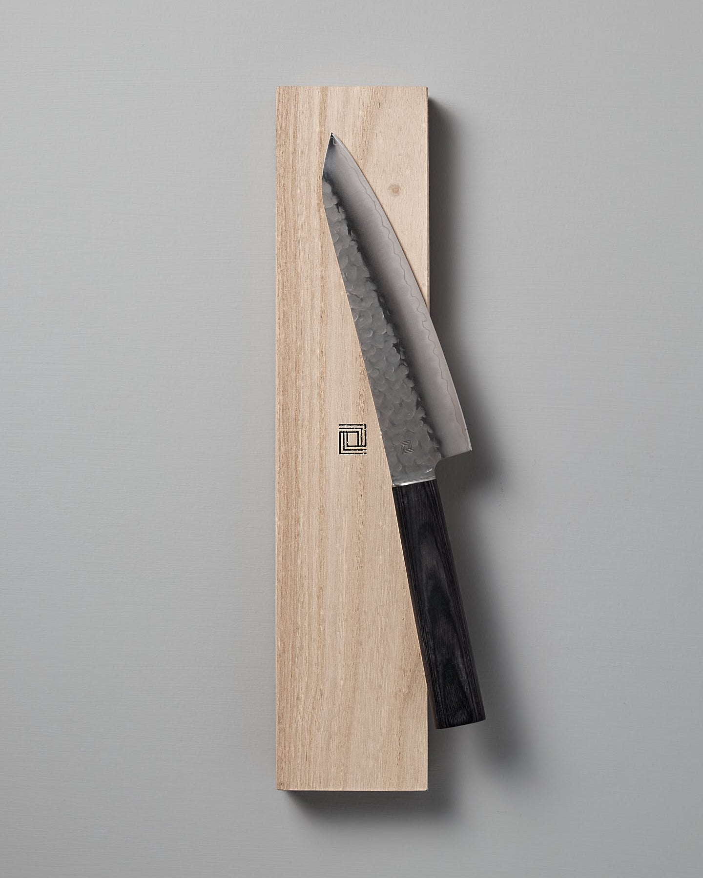 A Shizu Hamono Co. Yamato Santoku Knife with a dark handle rests on a rectangular wooden block against a plain gray background, showcasing the elegance of Japanese chef knives crafted from AUS8 stainless steel.