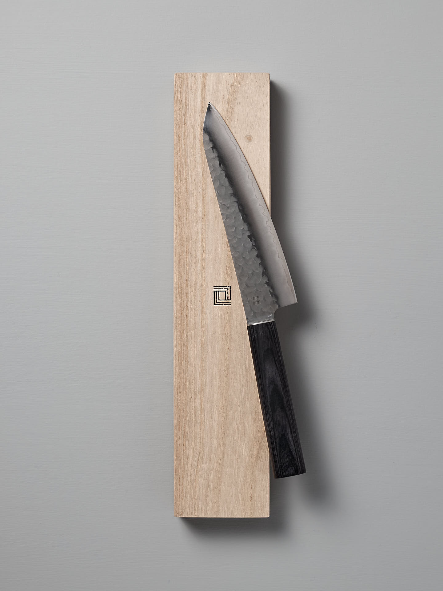 A Shizu Hamono Co. Yamato Santoku Knife with a dark handle rests on a rectangular wooden block against a plain gray background, showcasing the elegance of Japanese chef knives crafted from AUS8 stainless steel.