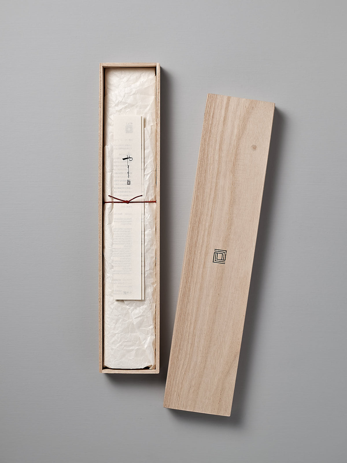 A long wooden box with the Shizu Hamono Co. logo on the lid, containing a rolled-up piece of paper tied with a red string, displayed on a light gray surface like those used to present the fine Yamato Santoku Knife.