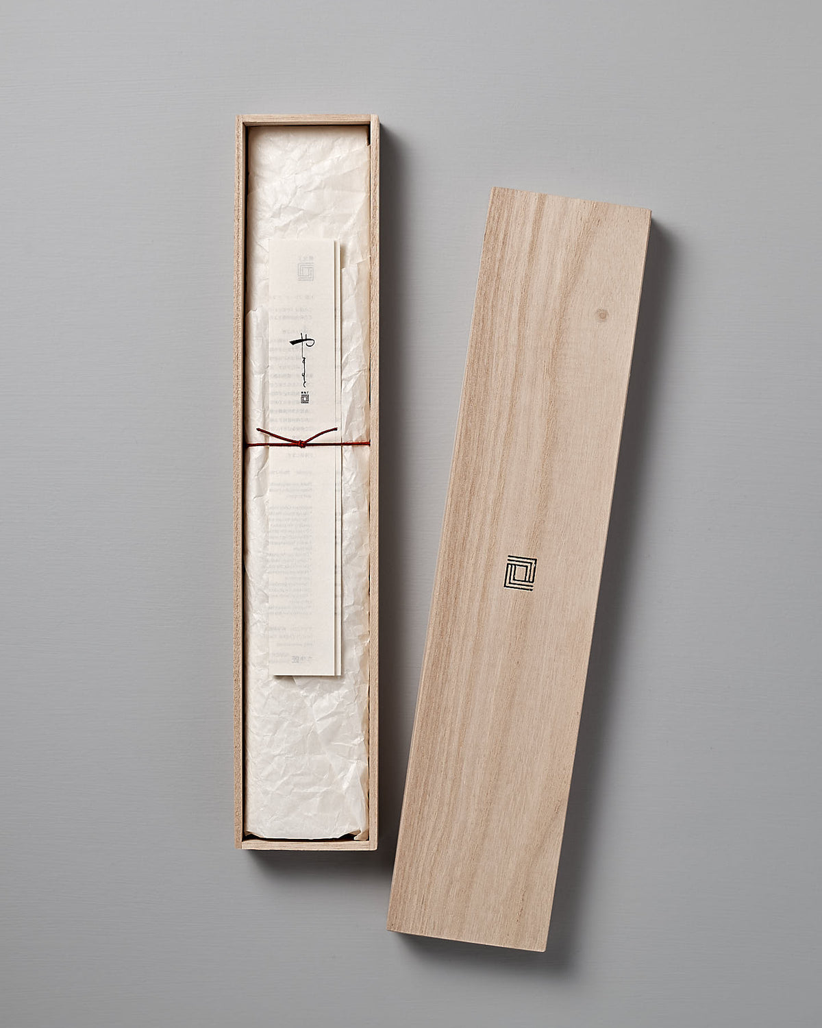 A long wooden box with the Shizu Hamono Co. logo on the lid, containing a rolled-up piece of paper tied with a red string, displayed on a light gray surface like those used to present the fine Yamato Santoku Knife.