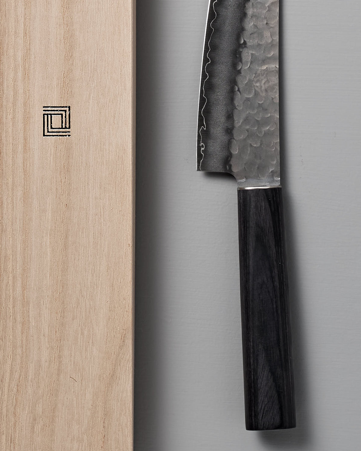 A close-up image of a Shizu Hamono Co. Yamato Santoku Knife with a textured AUS8 Japanese stainless steel blade and dark handle, positioned vertically next to a wooden block featuring a square logo.