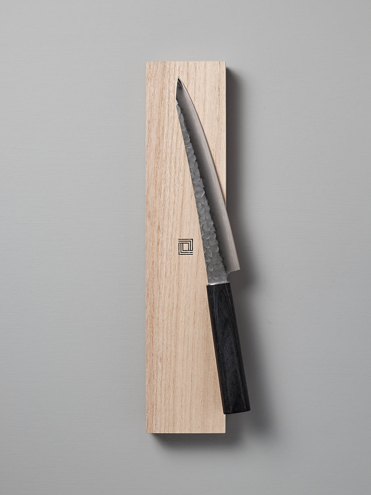 A Shizu Hamono Co. Yamato Yanagiba Knife with a black handle rests atop a wooden box adorned with a geometric logo on a light gray surface.