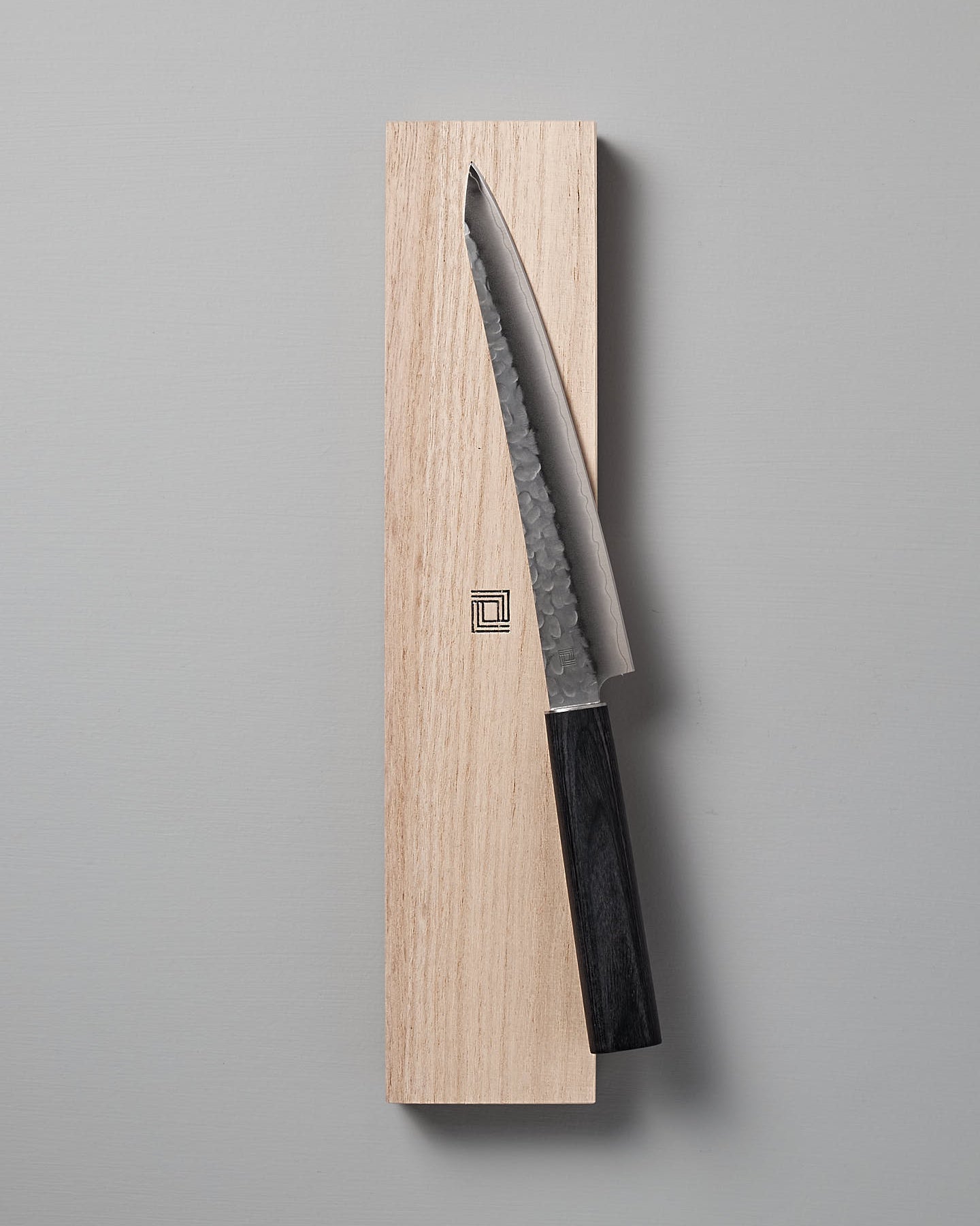 A Shizu Hamono Co. Yamato Yanagiba Knife with a black handle rests atop a wooden box adorned with a geometric logo on a light gray surface.