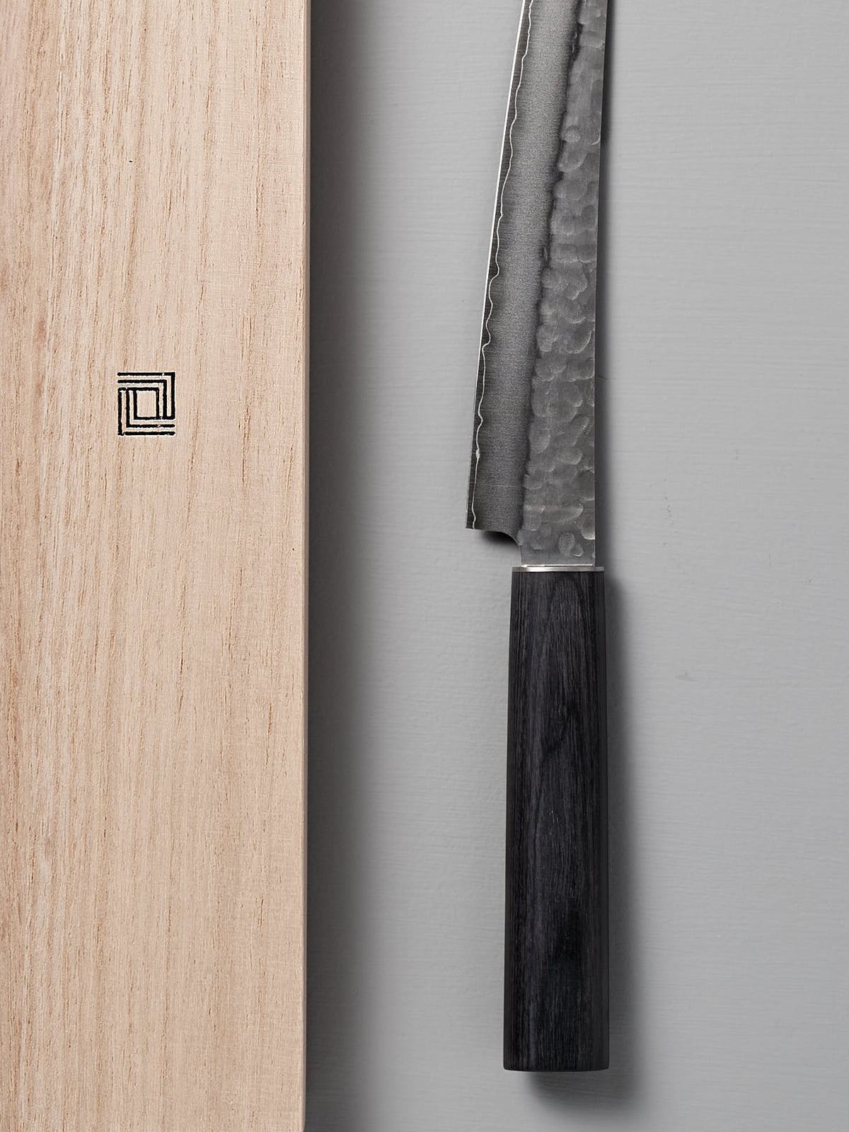 A close-up of the Yamato Yanagiba Knife by Shizu Hamono Co., showcasing its double-edged, hammered blade and wooden handle, positioned next to a wooden box adorned with a square geometric design.