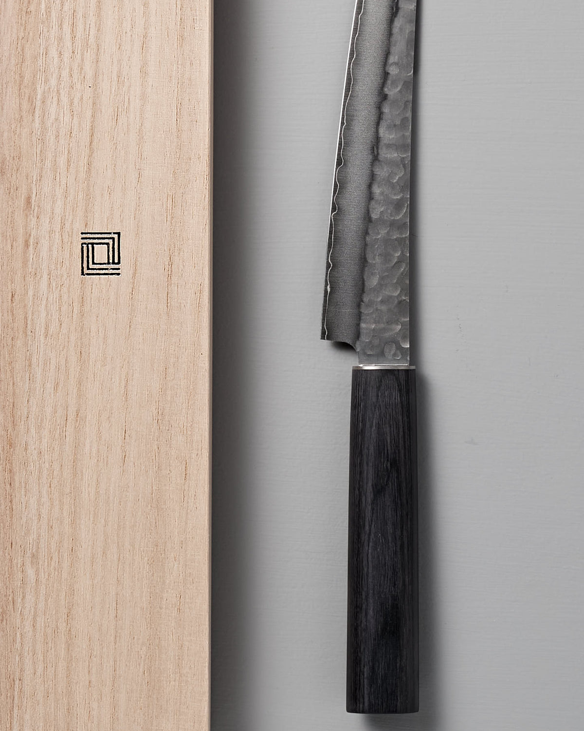 A close-up of the Yamato Yanagiba Knife by Shizu Hamono Co., showcasing its double-edged, hammered blade and wooden handle, positioned next to a wooden box adorned with a square geometric design.