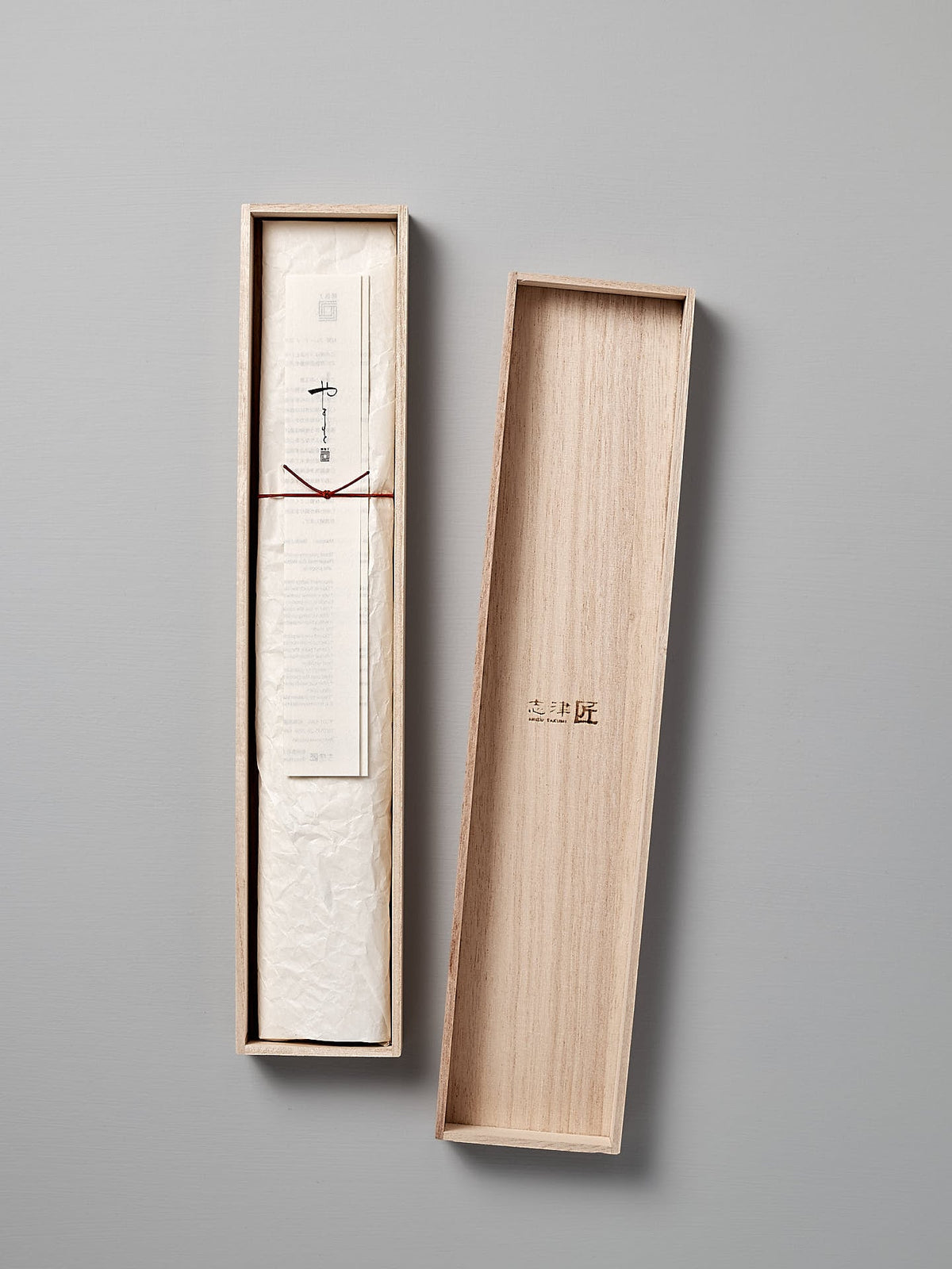 A piece of paper with Japanese text is neatly folded and placed inside an open wooden box with a lid resting on the side, reminiscent of the packaging for a fine Yamato Yanagiba Knife by Shizu Hamono Co.