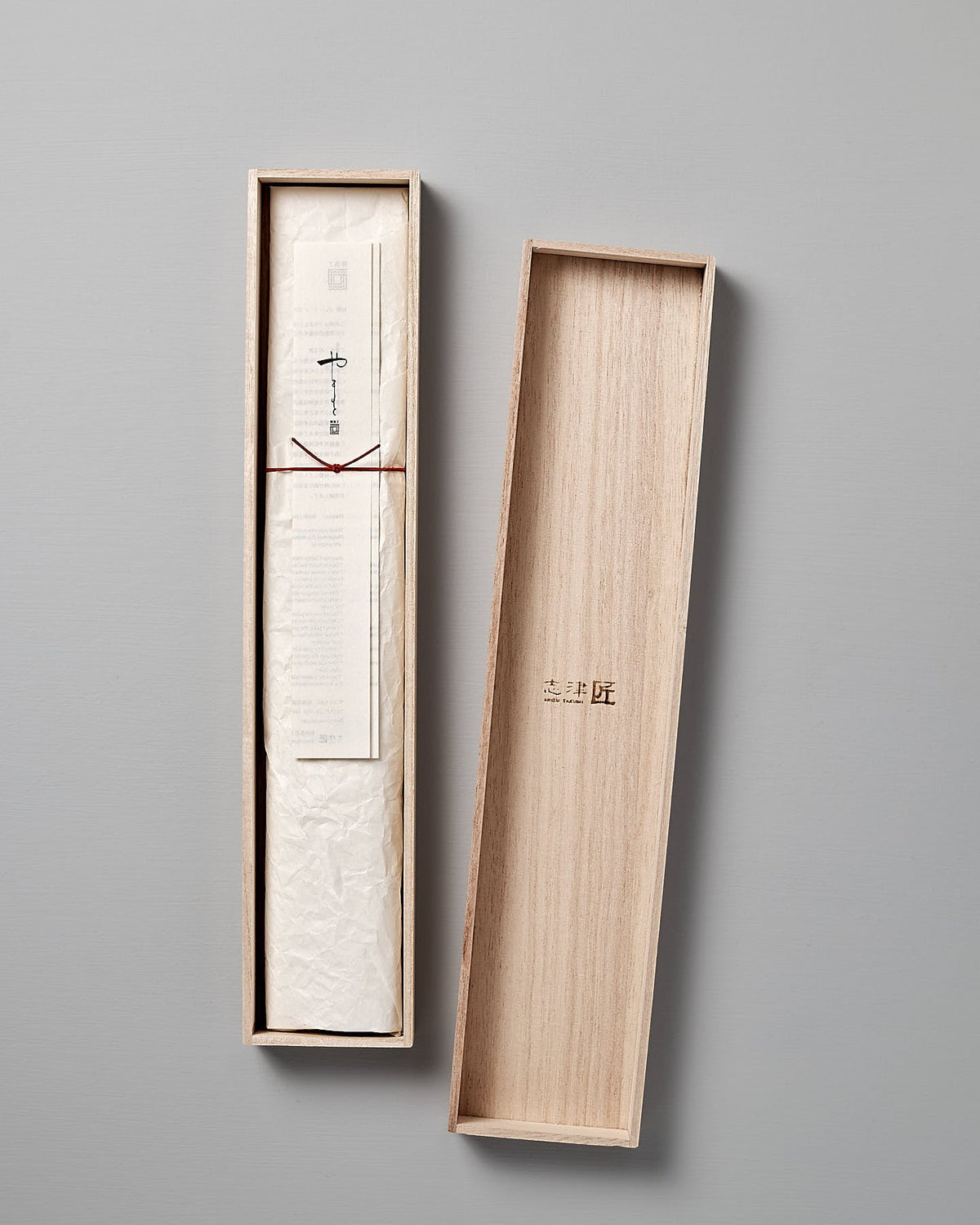 A piece of paper with Japanese text is neatly folded and placed inside an open wooden box with a lid resting on the side, reminiscent of the packaging for a fine Yamato Yanagiba Knife by Shizu Hamono Co.