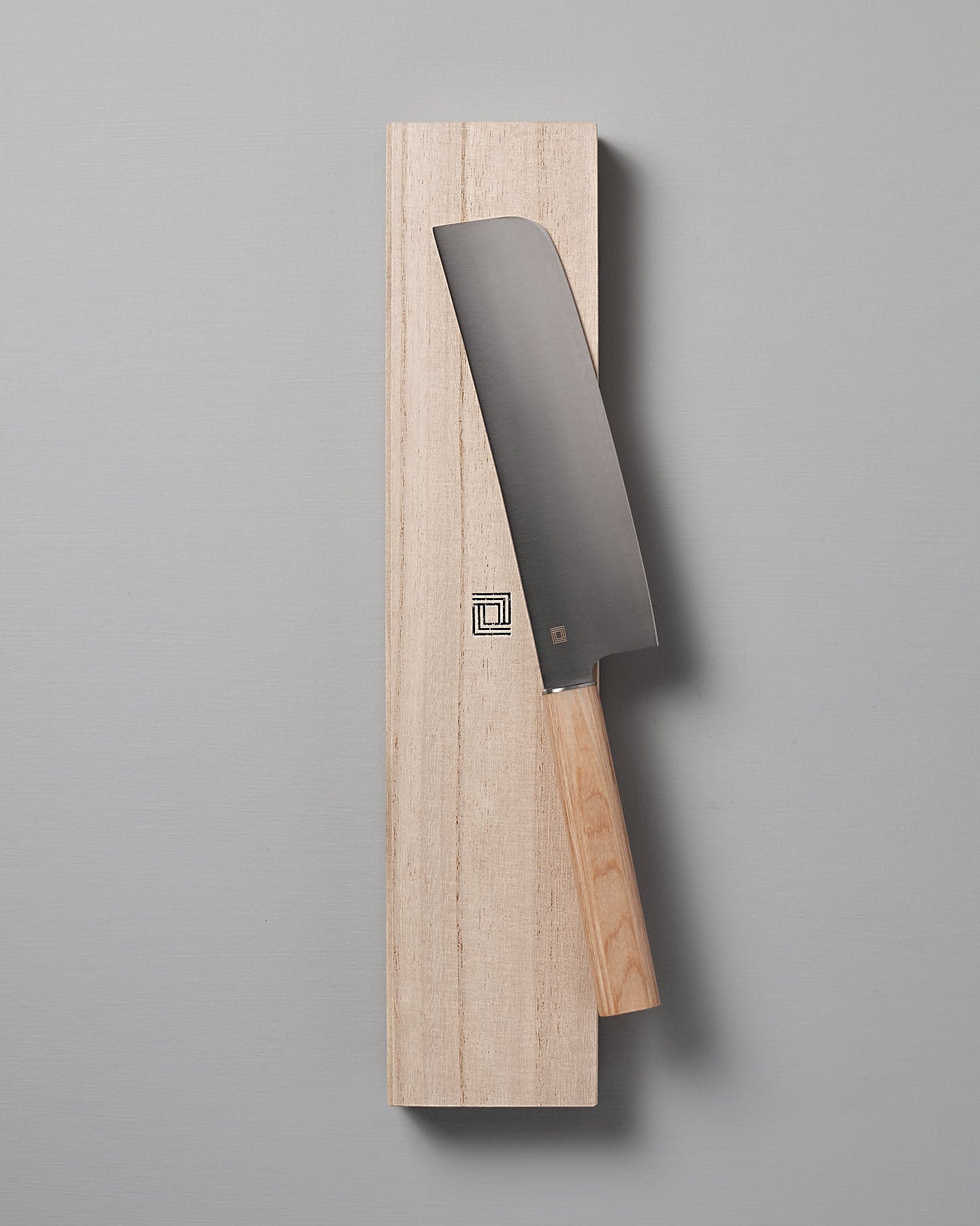 A Yuri Nakiri Knife from Shizu Hamono Co., featuring a thin blade and wooden handle, rests on a rectangular wooden block against a gray background, making it perfect for vegetable dicing.