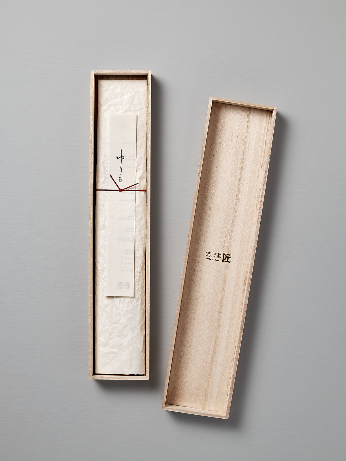 A slim, rectangular wooden box with an open lid reveals a rolled-up paper tied with a string inside. The box has minimalist markings and a smooth light-colored finish, subtly reminiscent of the elegance found in a Yuri Nakiri Knife by Shizu Hamono Co.