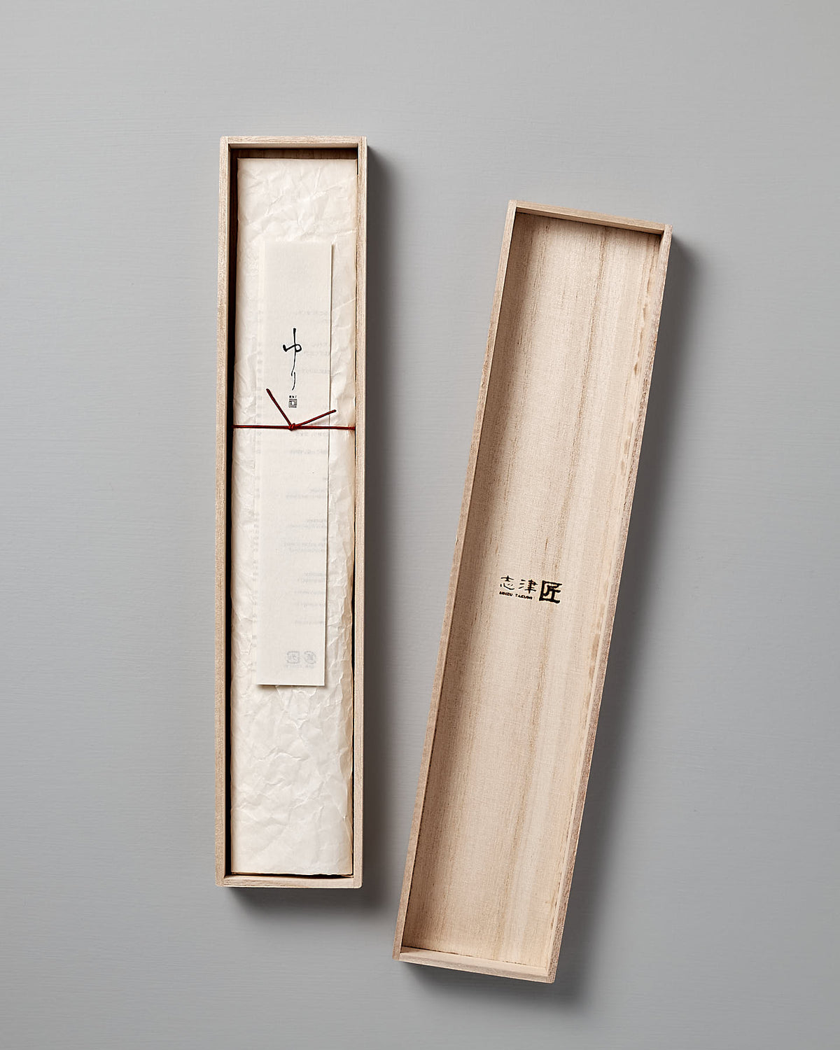 A slim, rectangular wooden box with an open lid reveals a rolled-up paper tied with a string inside. The box has minimalist markings and a smooth light-colored finish, subtly reminiscent of the elegance found in a Yuri Nakiri Knife by Shizu Hamono Co.
