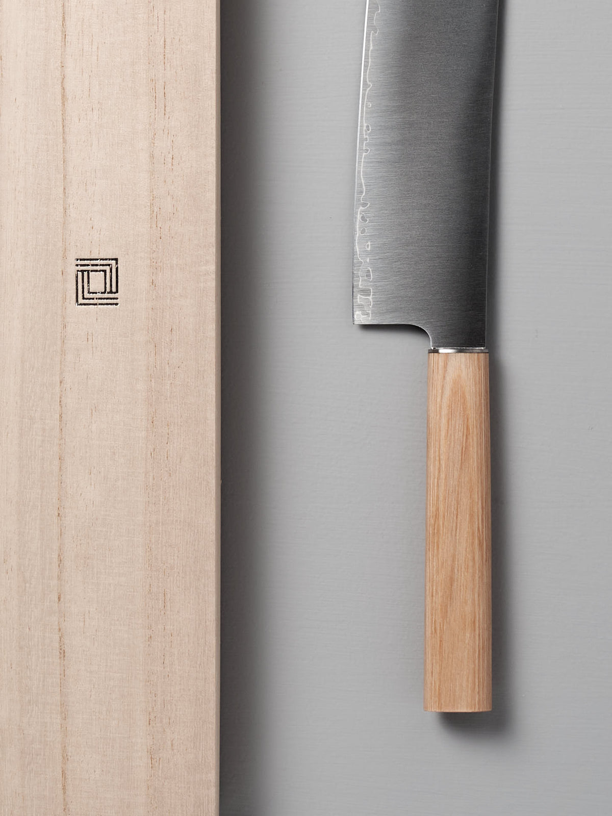 The Yuri Nakiri Knife by Shizu Hamono Co., featuring a wooden handle ideal for vegetable dicing, is displayed next to a wooden cutting board on a gray surface.