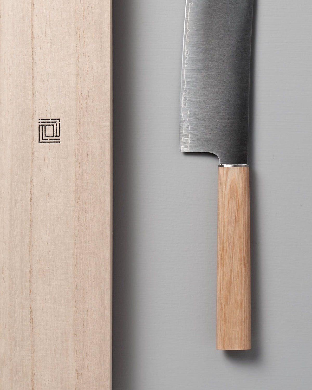 The Yuri Nakiri Knife by Shizu Hamono Co., featuring a wooden handle ideal for vegetable dicing, is displayed next to a wooden cutting board on a gray surface.