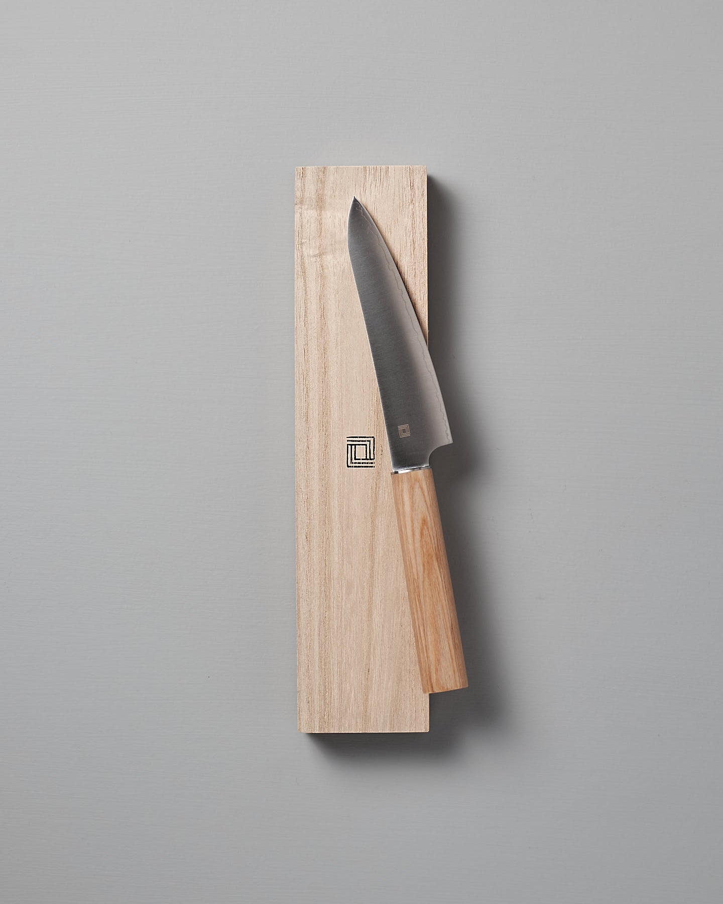 A Yuri Petite Knife from Shizu Hamono Co. with a wooden handle rests elegantly on a light wooden board against a gray background.