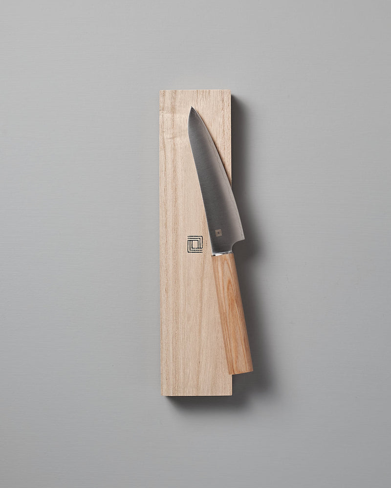 A Yuri Petite Knife from Shizu Hamono Co. with a wooden handle rests elegantly on a light wooden board against a gray background.