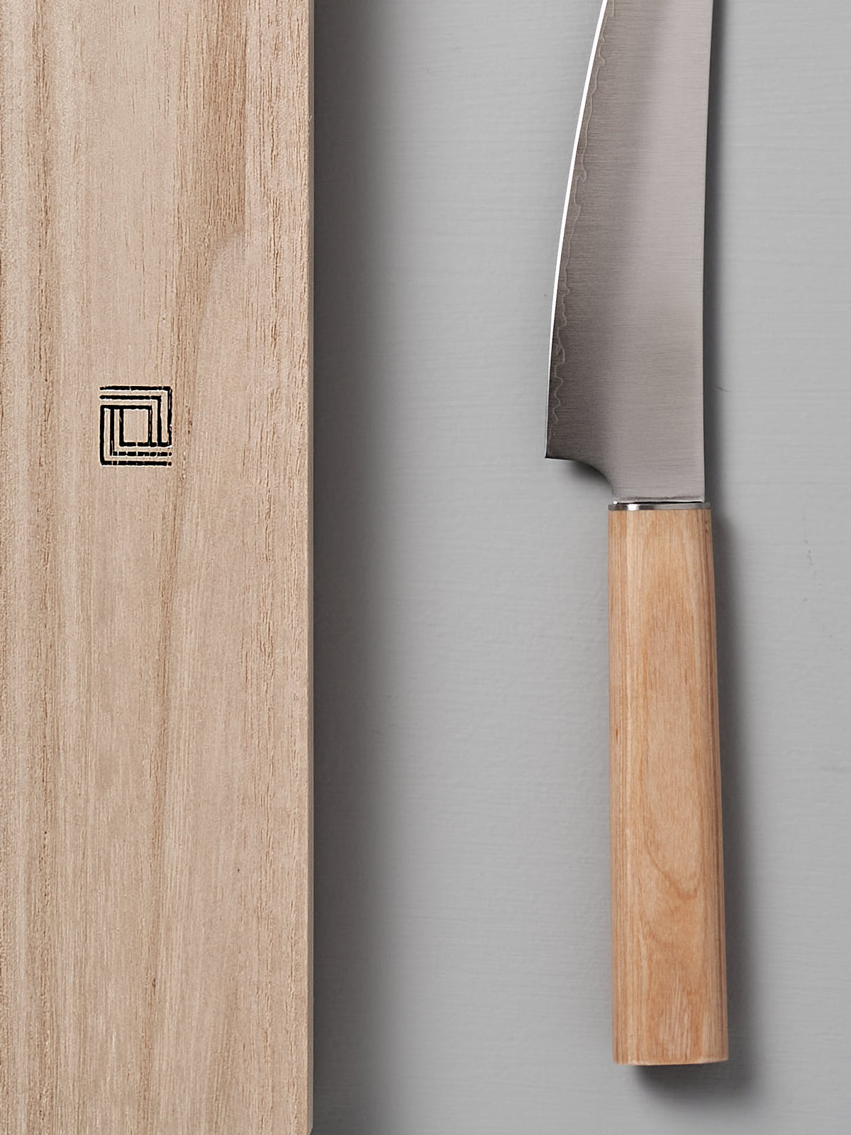 A Yuri Petite Knife from Shizu Hamono Co., featuring a wooden handle, lies beside a wooden cutting board with a geometric symbol on a gray background, exemplifying the precision and craftsmanship of Seki Japan.