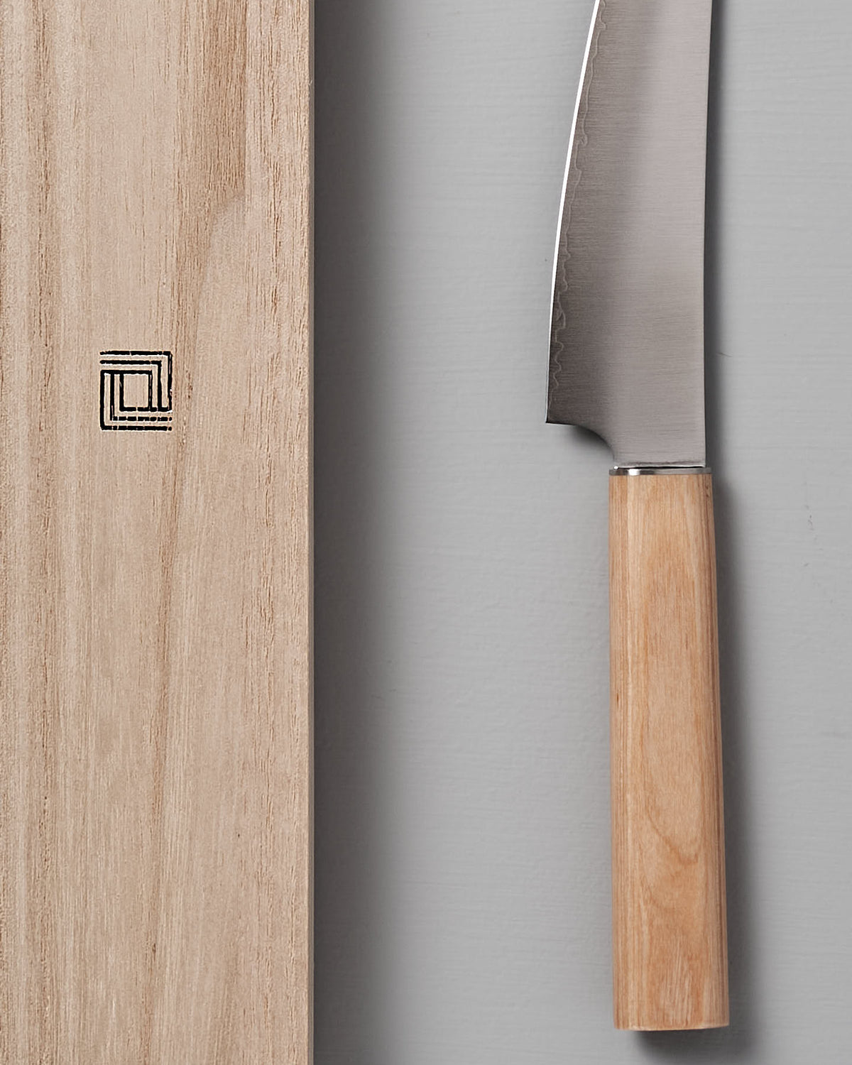 A Yuri Petite Knife from Shizu Hamono Co., featuring a wooden handle, lies beside a wooden cutting board with a geometric symbol on a gray background, exemplifying the precision and craftsmanship of Seki Japan.