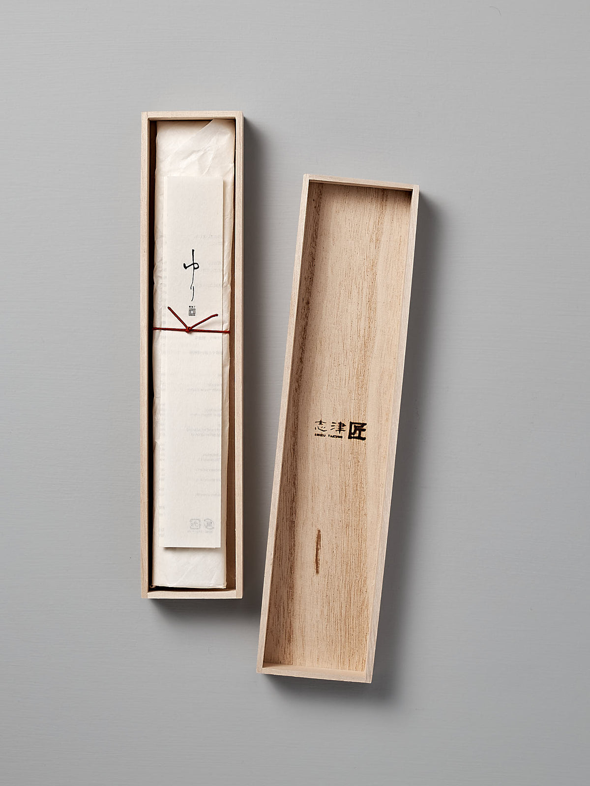 A wooden box with the lid removed, revealing a wrapped Yuri Petite Knife inside. The exterior lid features Japanese characters, hinting at its Seki Japan origin and its creation by Shizu Hamono Co. The background is a plain, light gray.