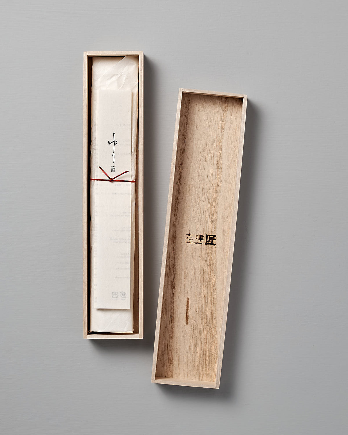 A wooden box with the lid removed, revealing a wrapped Yuri Petite Knife inside. The exterior lid features Japanese characters, hinting at its Seki Japan origin and its creation by Shizu Hamono Co. The background is a plain, light gray.