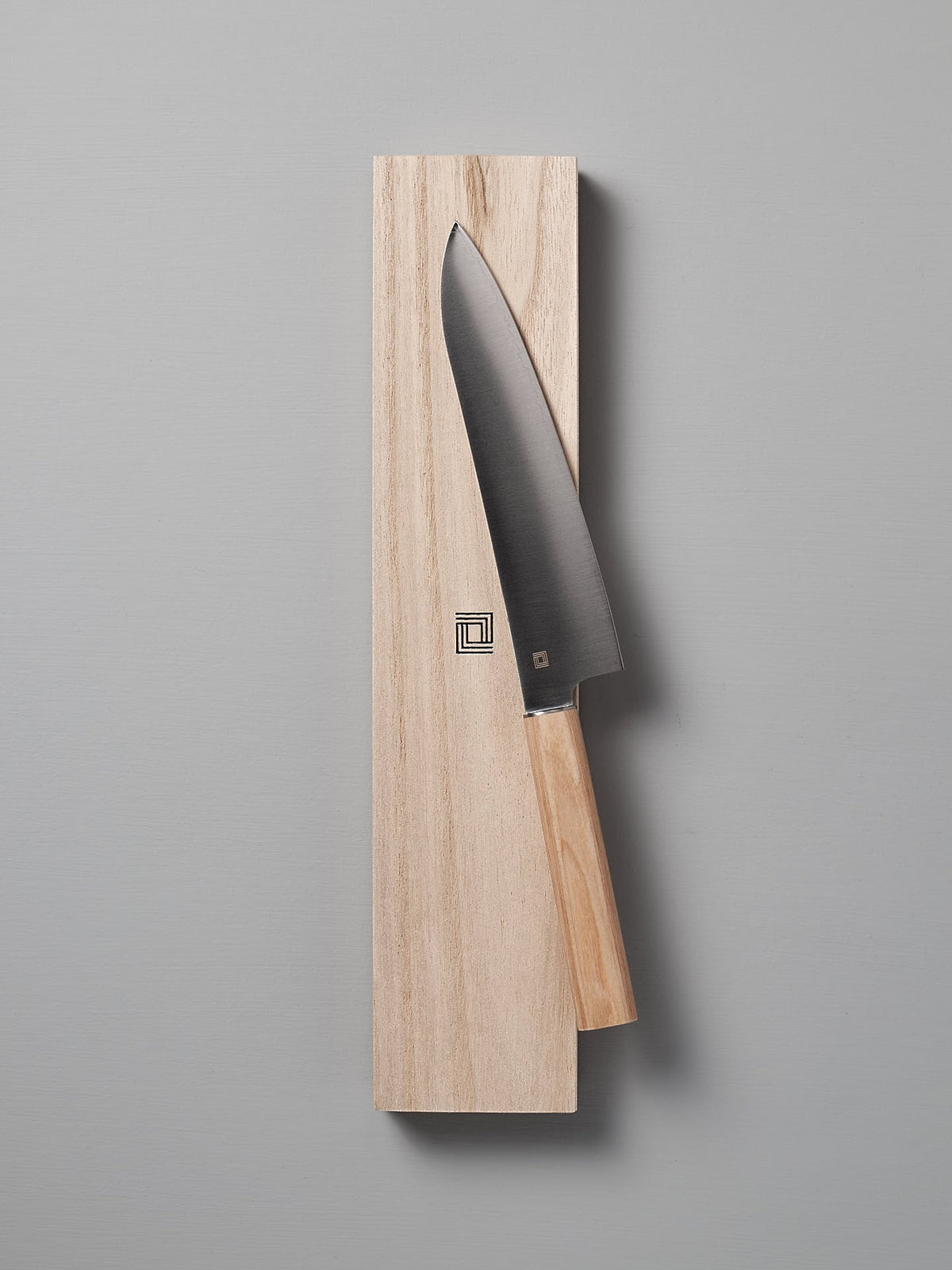 A Yuri Santoku Knife by Shizu Hamono Co., featuring a light wooden handle, is elegantly placed on a rectangular wooden block against a plain gray background, highlighting the superior craftsmanship of Japanese Chef knives.