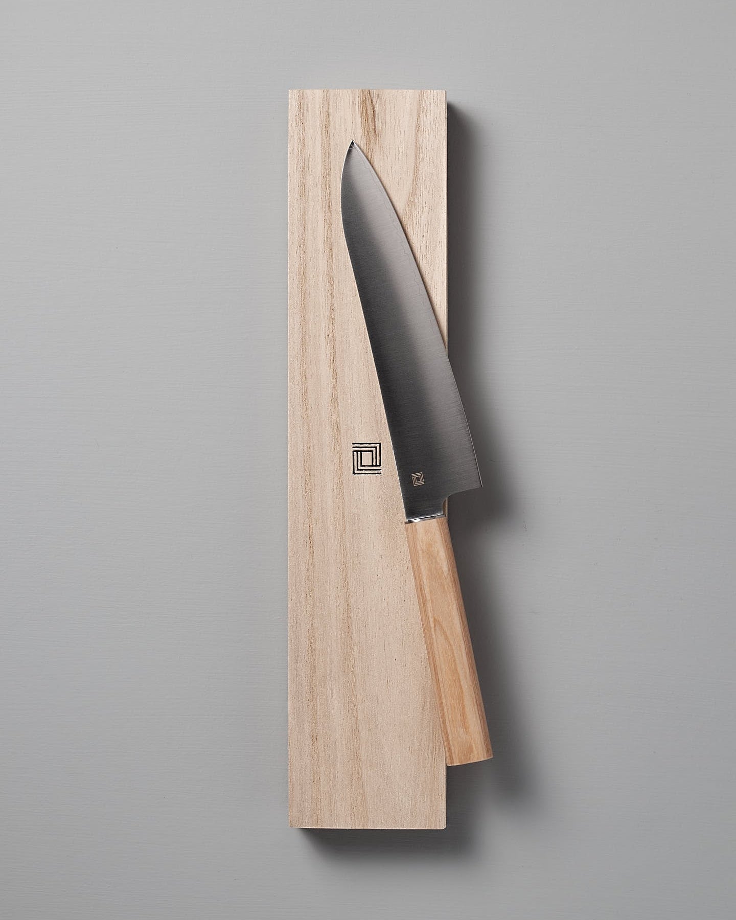 A Yuri Santoku Knife by Shizu Hamono Co., featuring a light wooden handle, is elegantly placed on a rectangular wooden block against a plain gray background, highlighting the superior craftsmanship of Japanese Chef knives.