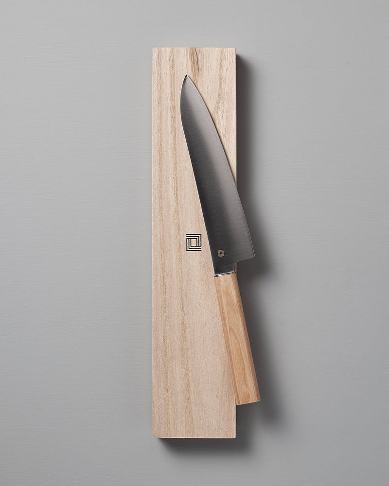 A Yuri Santoku Knife by Shizu Hamono Co., featuring a light wooden handle, is elegantly placed on a rectangular wooden block against a plain gray background, highlighting the superior craftsmanship of Japanese Chef knives.