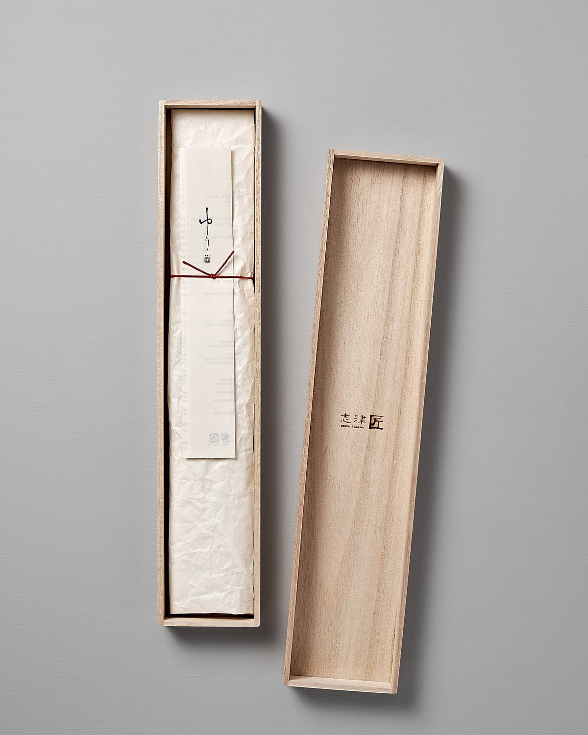 A wooden box with a lid reveals a neatly wrapped paper package and a small tag with text inside on a gray background, hinting at the exquisite Yuri Santoku Knife by Shizu Hamono Co. within.