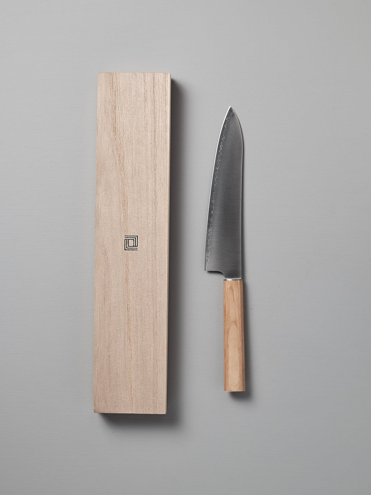 A Shizu Hamono Co. Yuri Santoku Knife, featuring a stainless steel blade and a wooden handle, is placed beside its matching wooden sheath on a grey surface.