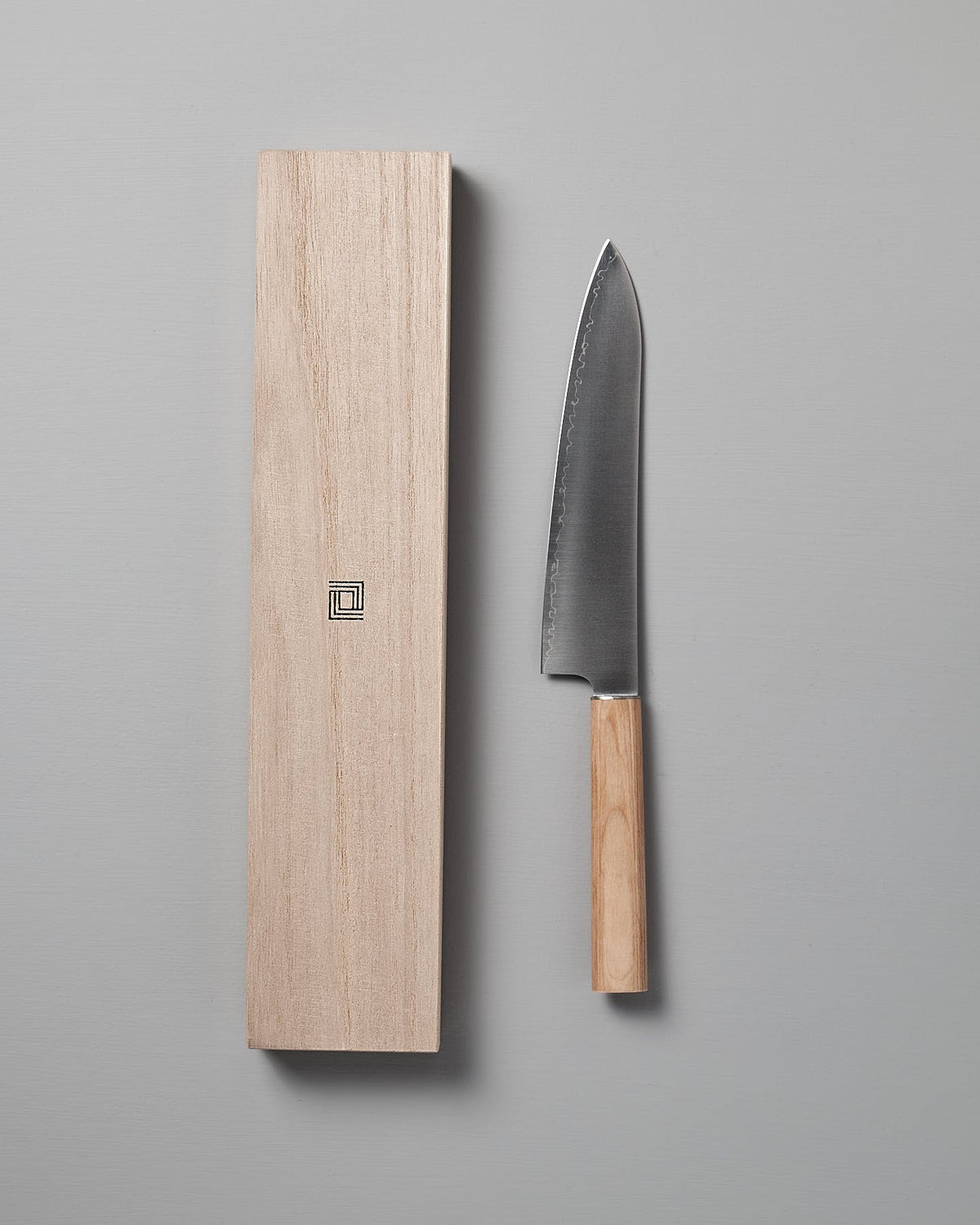 A Shizu Hamono Co. Yuri Santoku Knife, featuring a stainless steel blade and a wooden handle, is placed beside its matching wooden sheath on a grey surface.