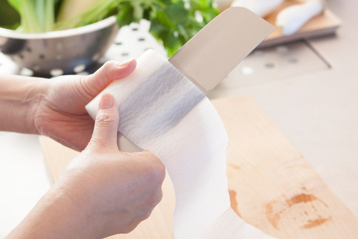 A person wiping a stainless steel blade of a Yuri Nakiri Knife by Shizu Hamono Co. with a white cloth in a kitchen setting, while a cutting board and a colander filled with green vegetables are visible in the background.