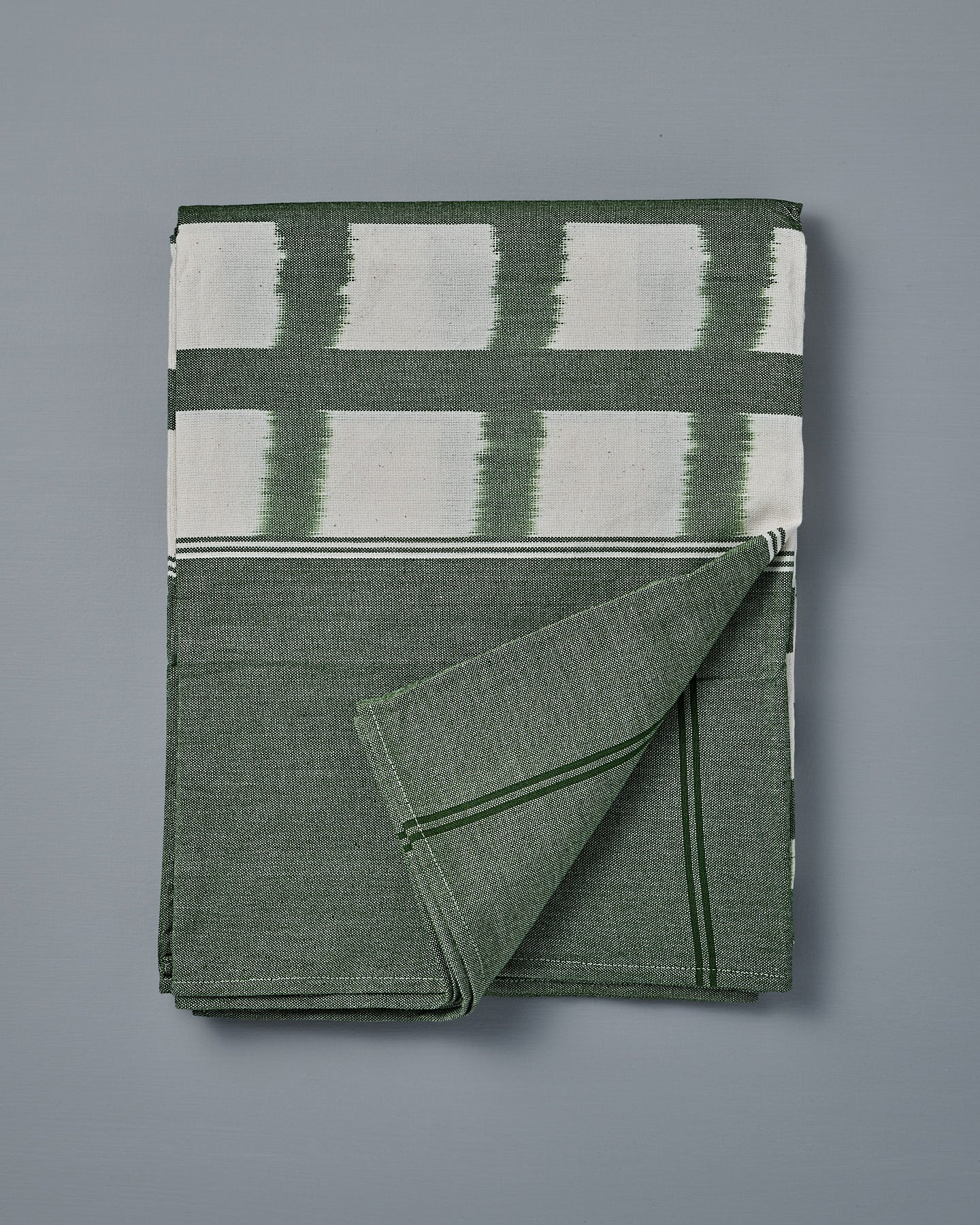 The Ikat Weave Tablecloth – Green Grid, made in India with a green and white design, is shown folded on a flat gray surface with one corner turned up.