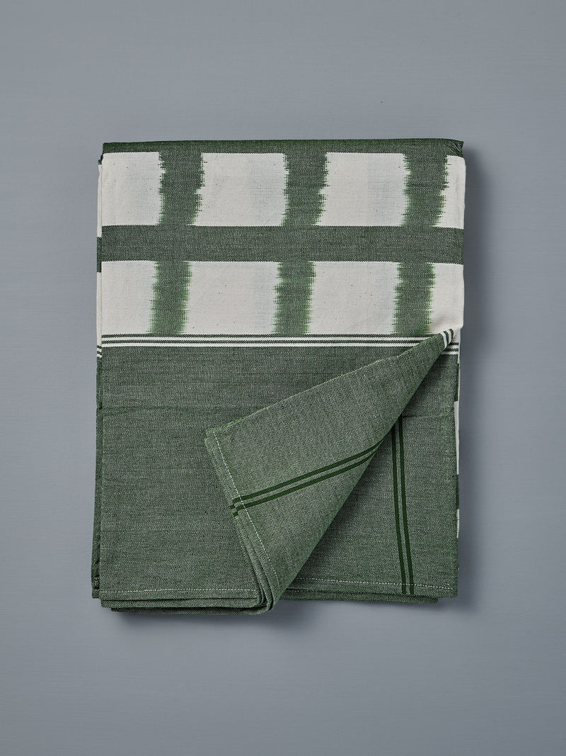 The Ikat Weave Tablecloth – Green Grid, made in India with a green and white design, is shown folded on a flat gray surface with one corner turned up.