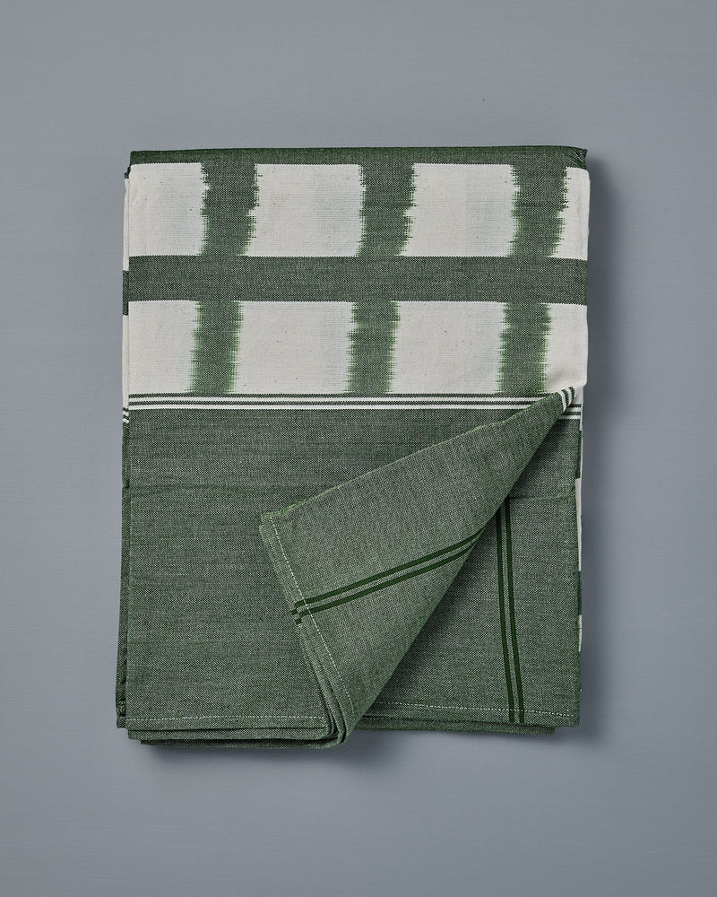 The Ikat Weave Tablecloth – Green Grid, made in India with a green and white design, is shown folded on a flat gray surface with one corner turned up.