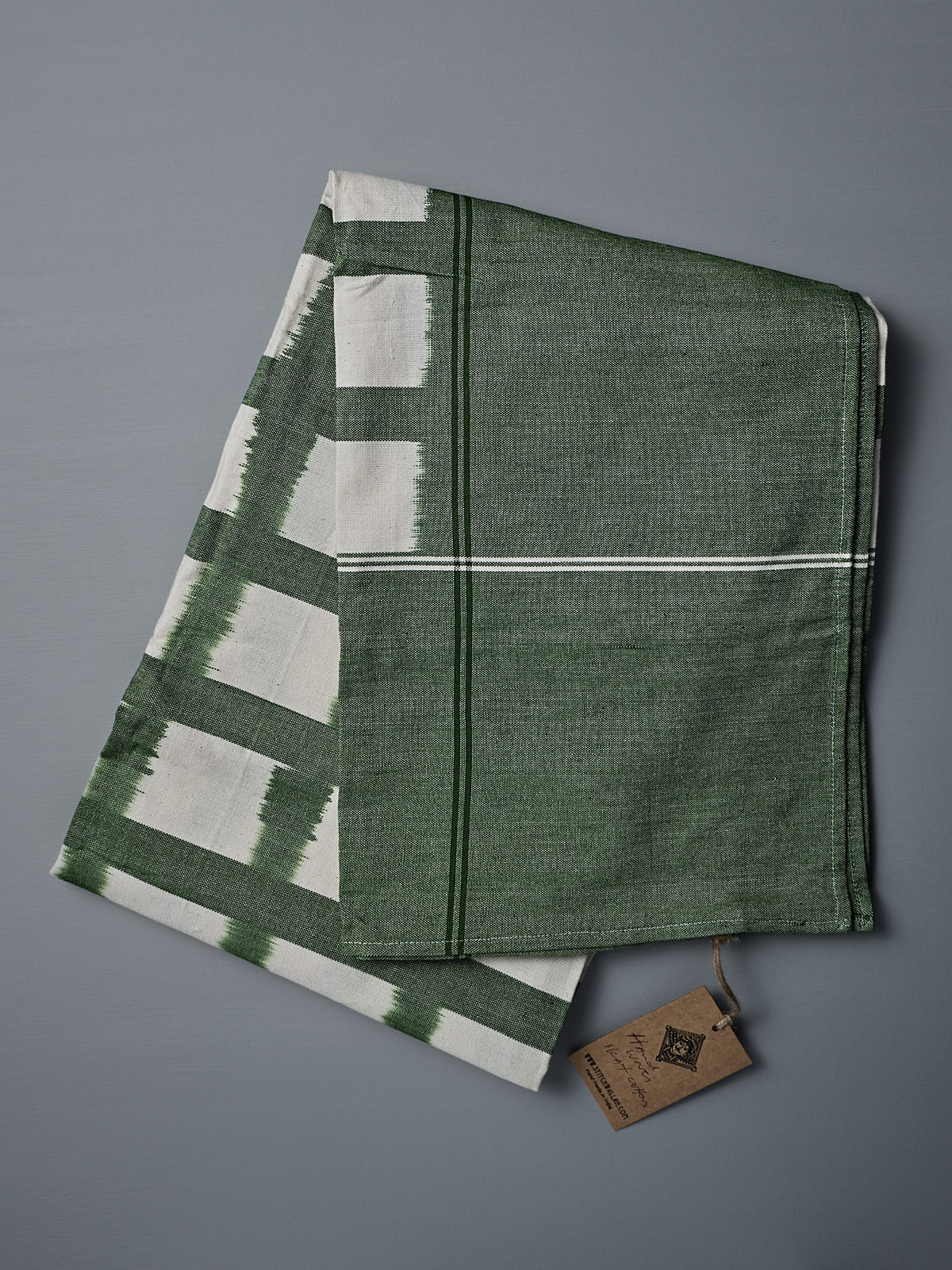 A folded green and white Ikat Weave Tablecloth – Green Grid, with a tag attached, is placed on a gray surface.