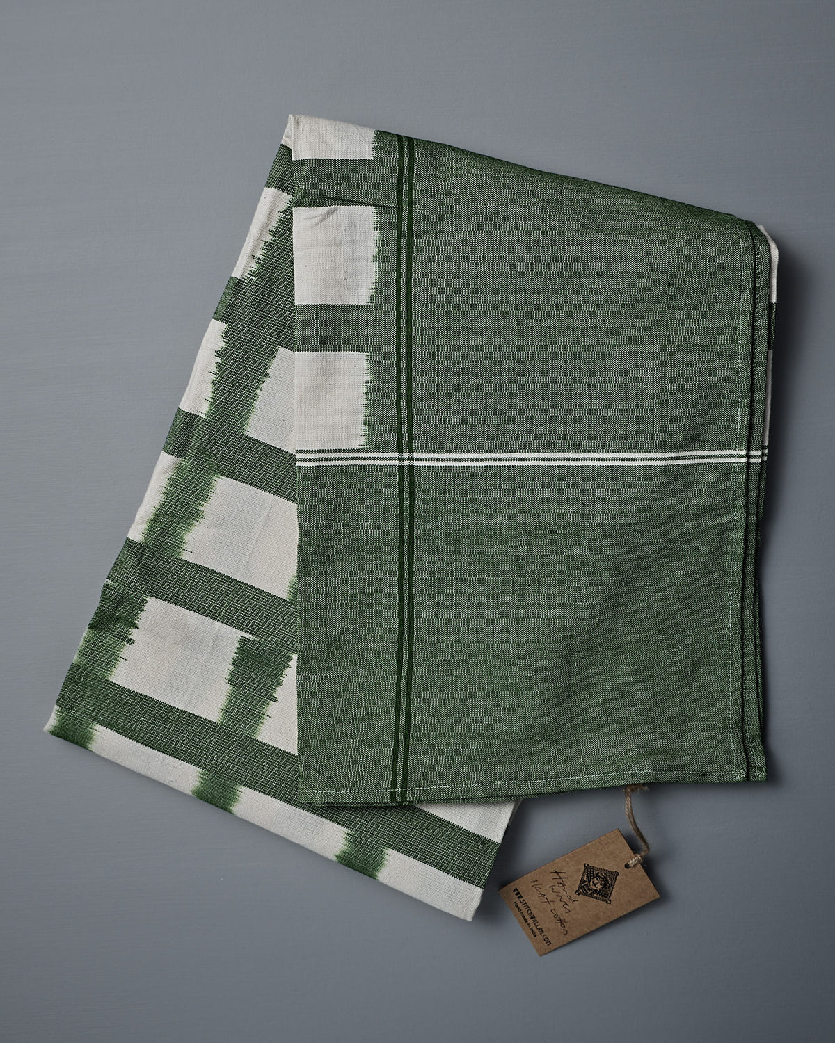 A folded green and white Ikat Weave Tablecloth – Green Grid, with a tag attached, is placed on a gray surface.