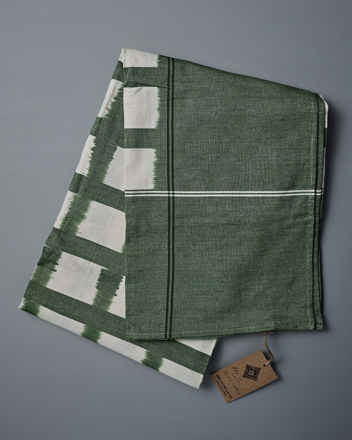 The Ikat Weave Tablecloth – Green Grid, made in India with a green and white design, is shown folded on a flat gray surface with one corner turned up.