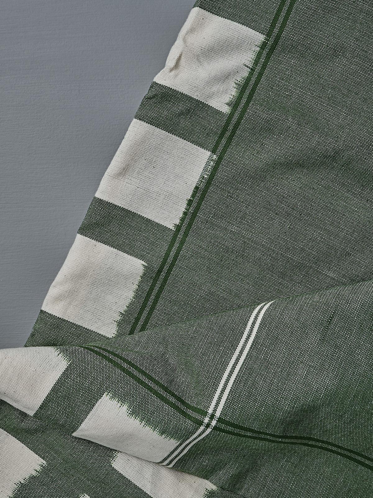 A close-up of the Ikat Weave Tablecloth – Green Grid, a handwoven green and white cotton fabric with geometric patterns, laid on a gray surface—crafted in India.