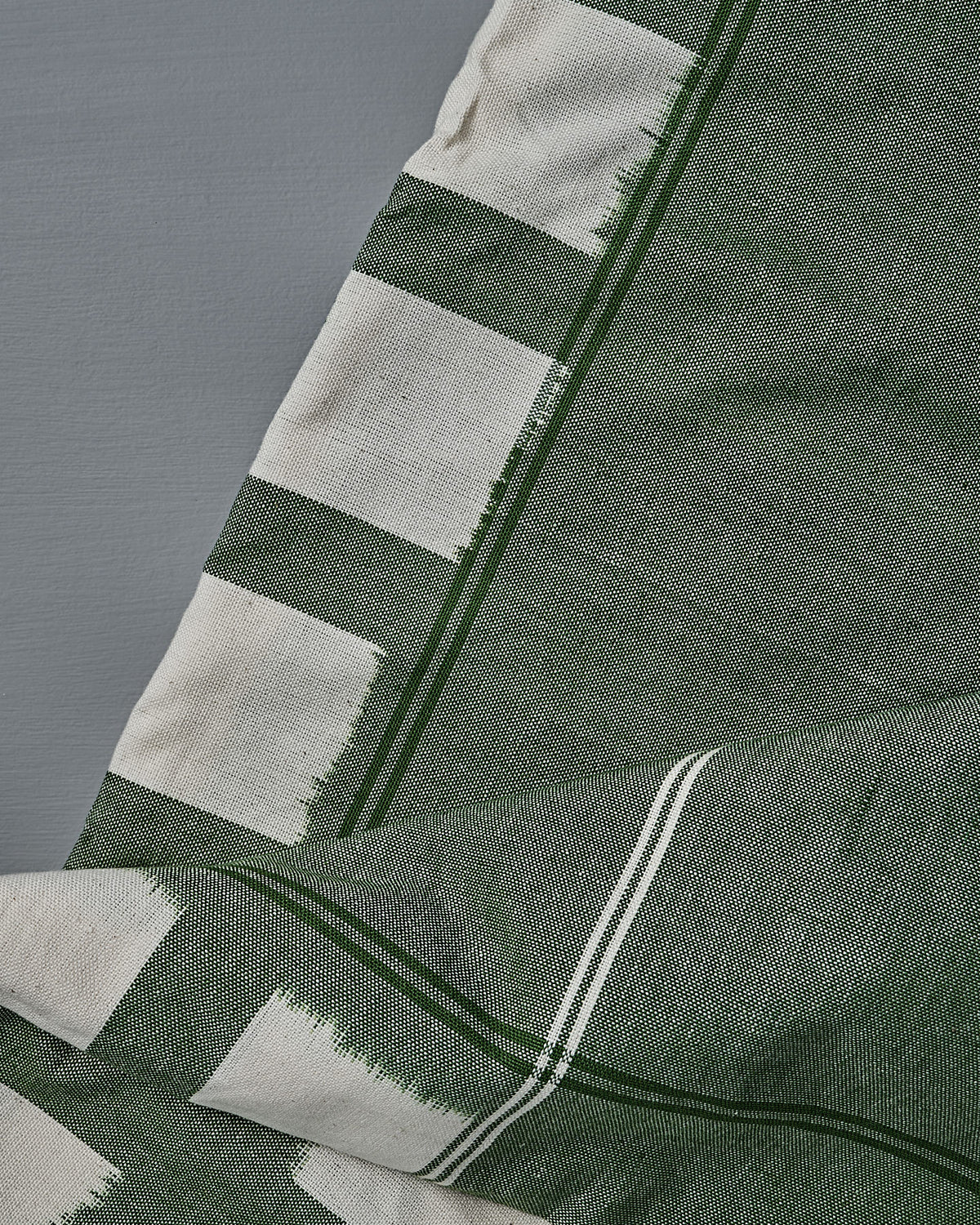 A close-up of the Ikat Weave Tablecloth – Green Grid, a handwoven green and white cotton fabric with geometric patterns, laid on a gray surface—crafted in India.