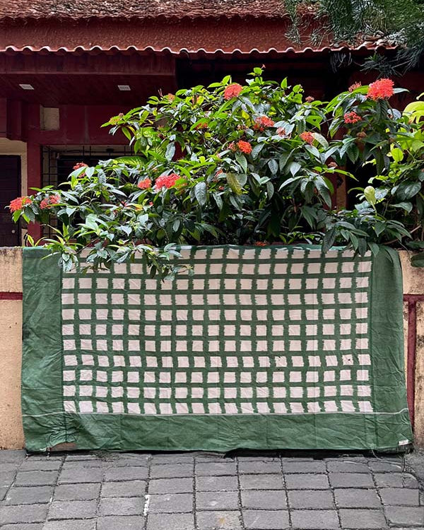 The Ikat Weave Tablecloth – Green Grid, a handwoven cotton piece in green and white checks, is draped over a low wall adorned with leafy plants and red flowers, set before a red building.