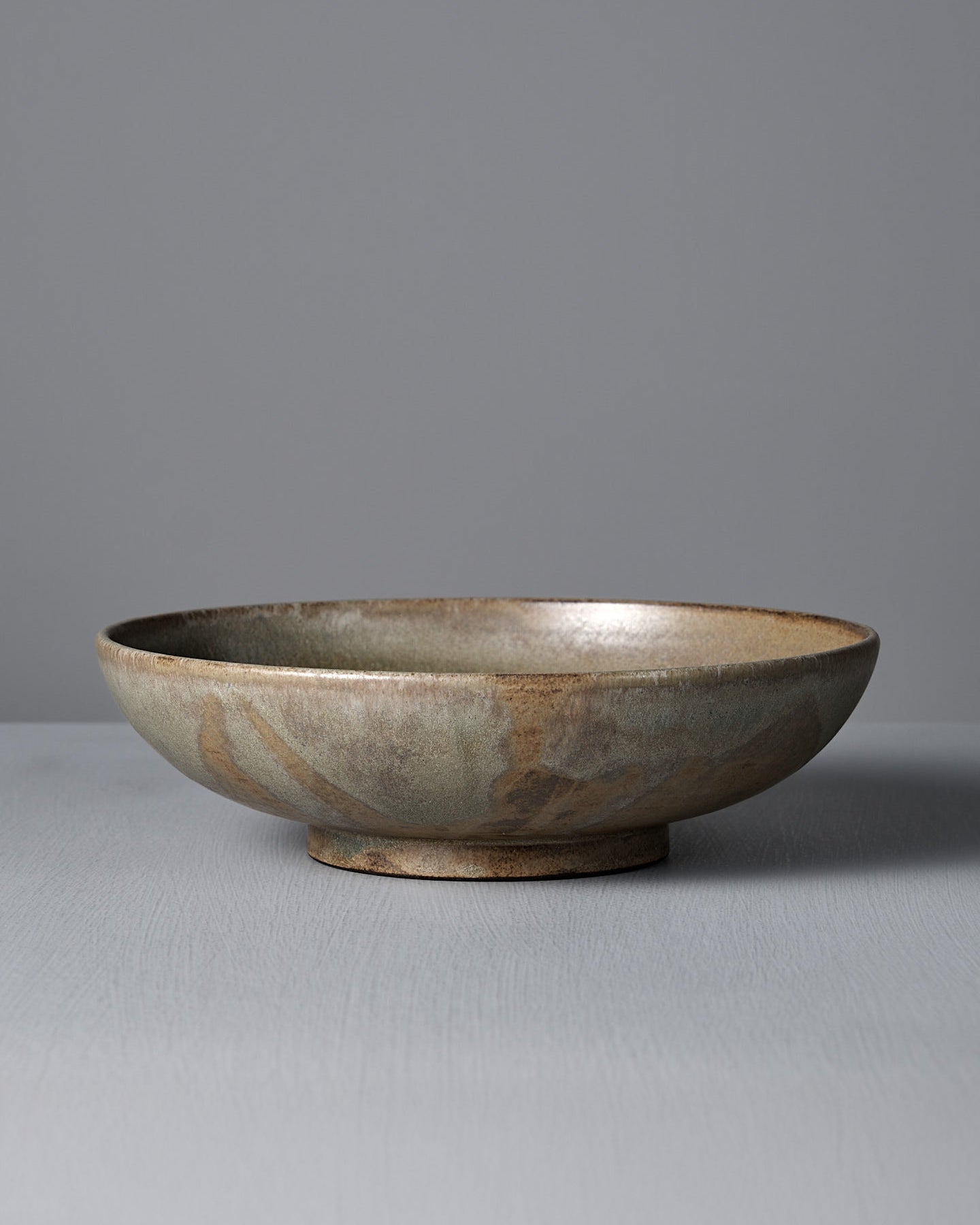 The Studio Foon Shallow Serving Bowl – Raw Umber, a handmade round bowl with a mottled brown and gray glaze, sits on a flat light gray surface against a plain gray background.