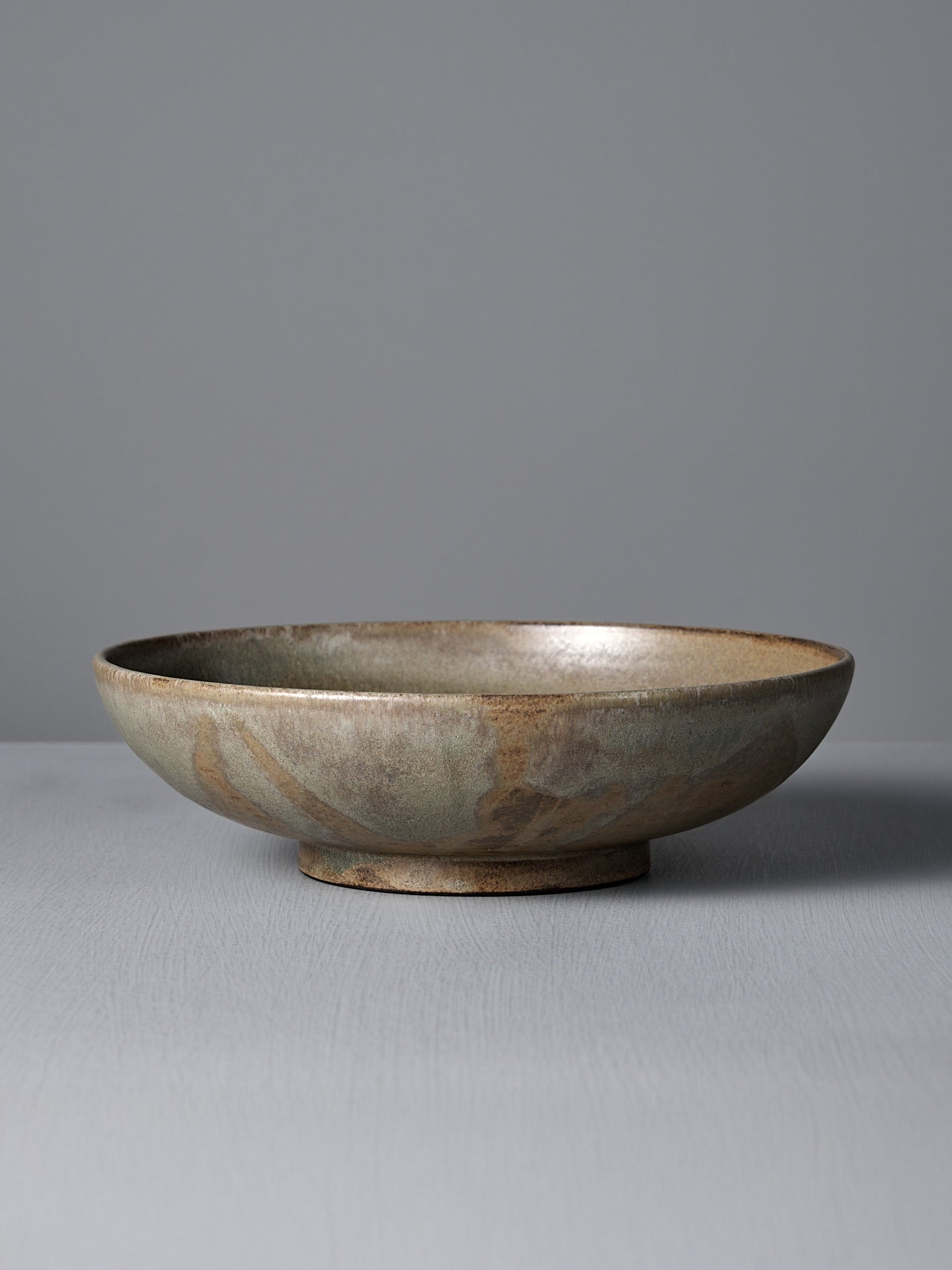 The Studio Foon Shallow Serving Bowl – Raw Umber, a handmade round bowl with a mottled brown and gray glaze, sits on a flat light gray surface against a plain gray background.