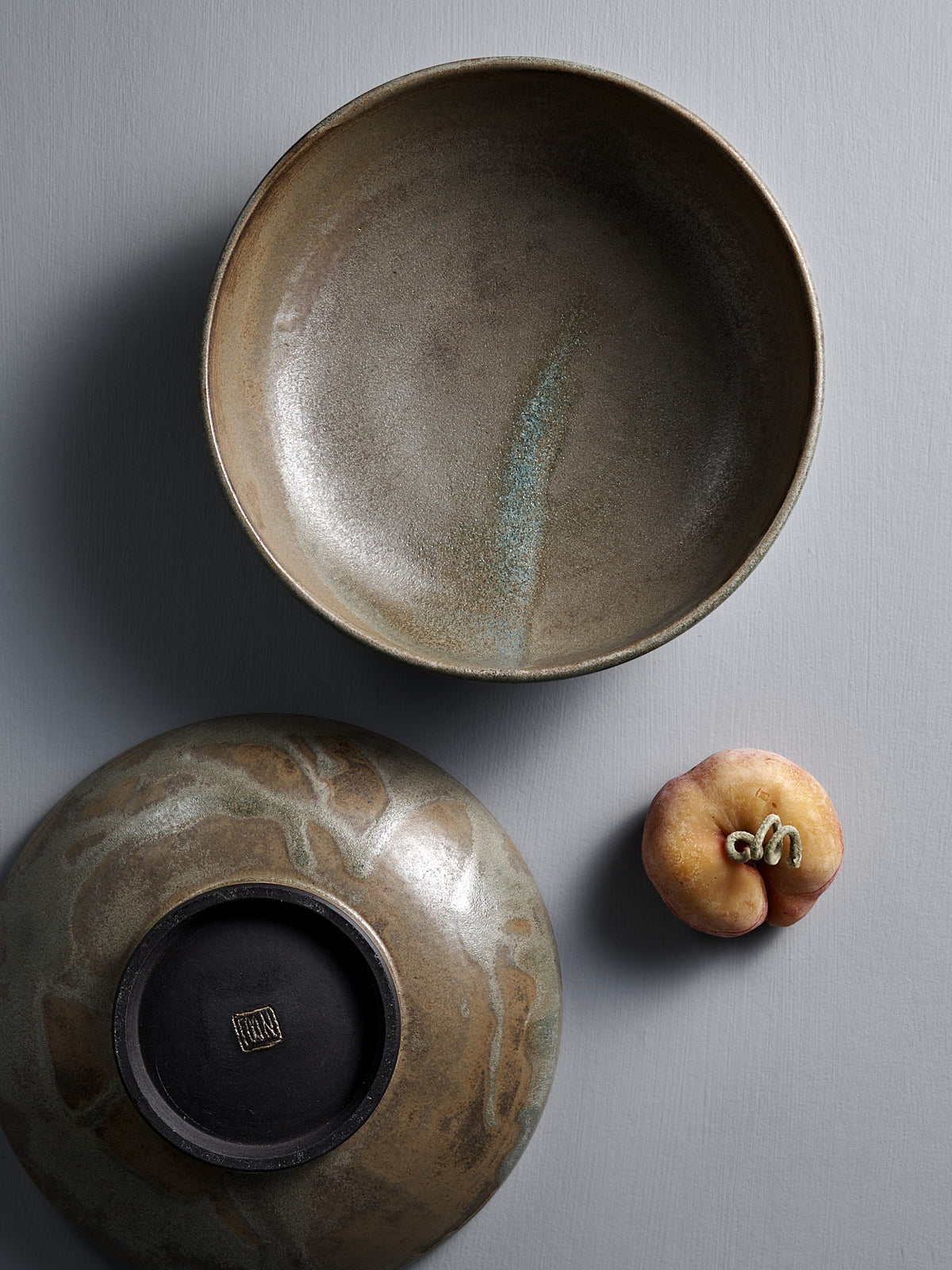 A Studio Foon Shallow Serving Bowl in Raw Umber, another bowl displayed upside down, and a flat peach are arranged on a gray textured surface.