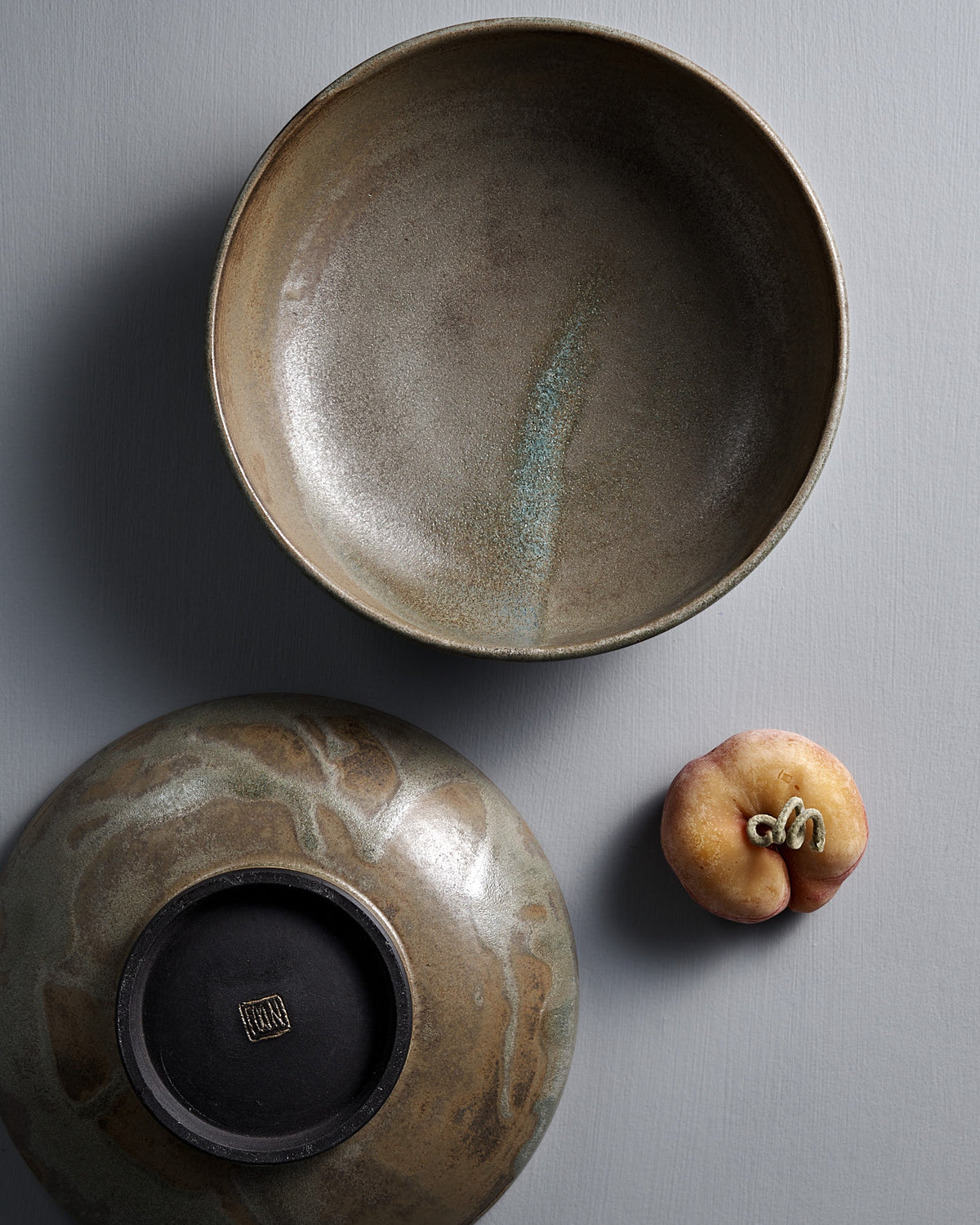 A Studio Foon Shallow Serving Bowl in Raw Umber, another bowl displayed upside down, and a flat peach are arranged on a gray textured surface.