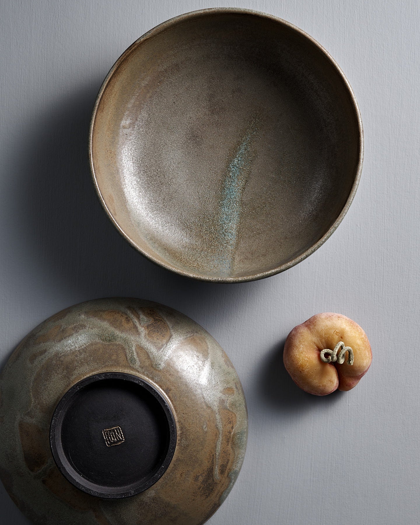 The Studio Foon Shallow Serving Bowl – Raw Umber, a handmade round bowl with a mottled brown and gray glaze, sits on a flat light gray surface against a plain gray background.