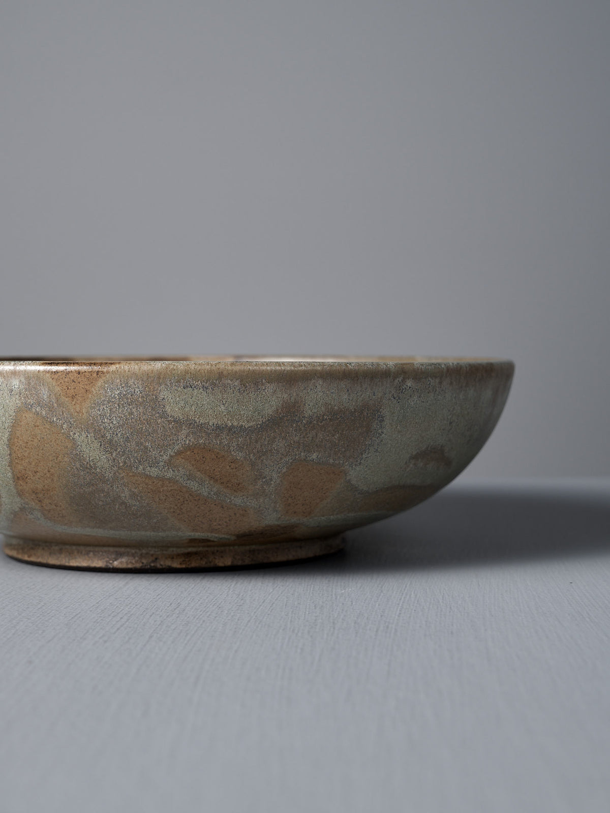 The Shallow Serving Bowl – Raw Umber by Studio Foon, featuring a matte earth-toned glaze, sits on a gray surface with a plain gray background.