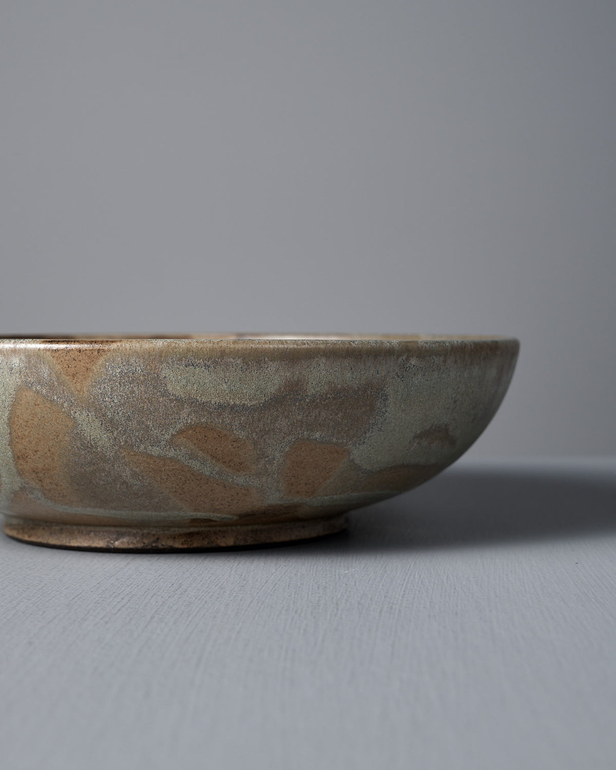 The Shallow Serving Bowl – Raw Umber by Studio Foon, featuring a matte earth-toned glaze, sits on a gray surface with a plain gray background.