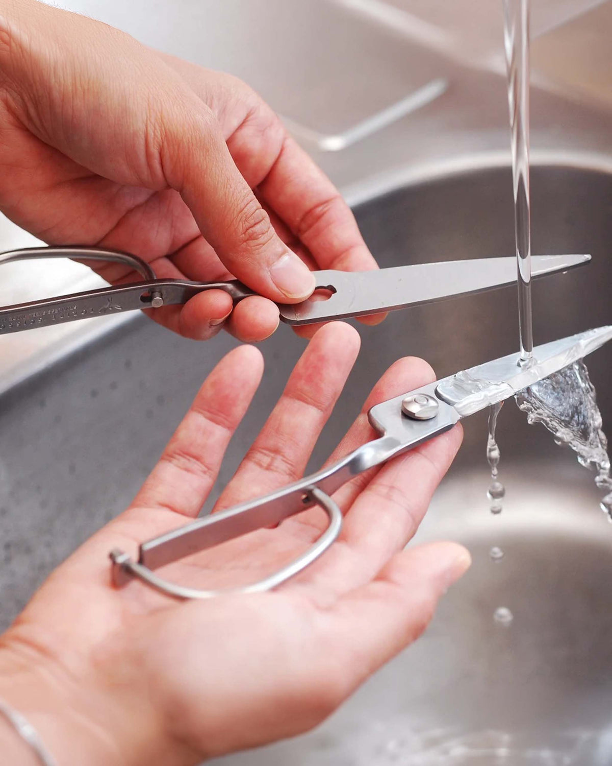 Hands washing a pair of metal kitchen shears under running water in a sink, with the scissors taken apart into two separate pieces.