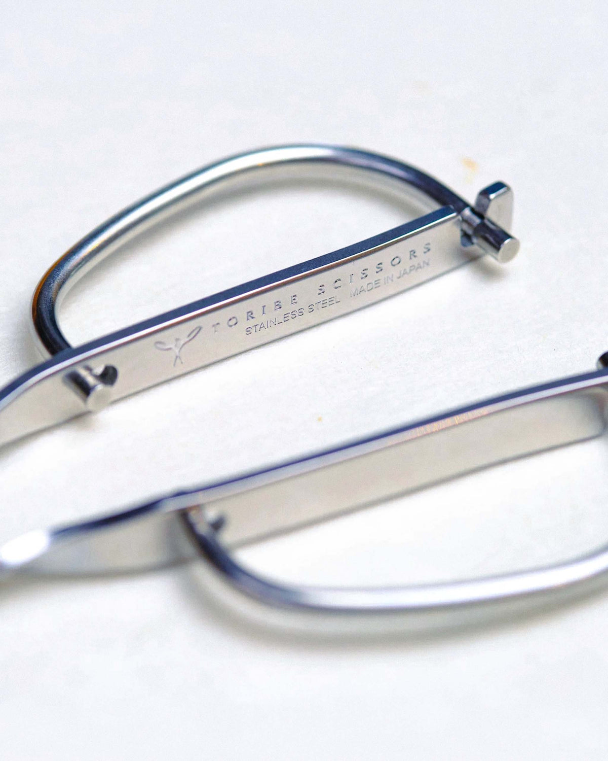 Close-up of two stainless steel eyebrow scissors with flat handles, labeled “TORIBE SCISSORS STAINLESS STEEL MADE IN JAPAN,” placed on a white surface.